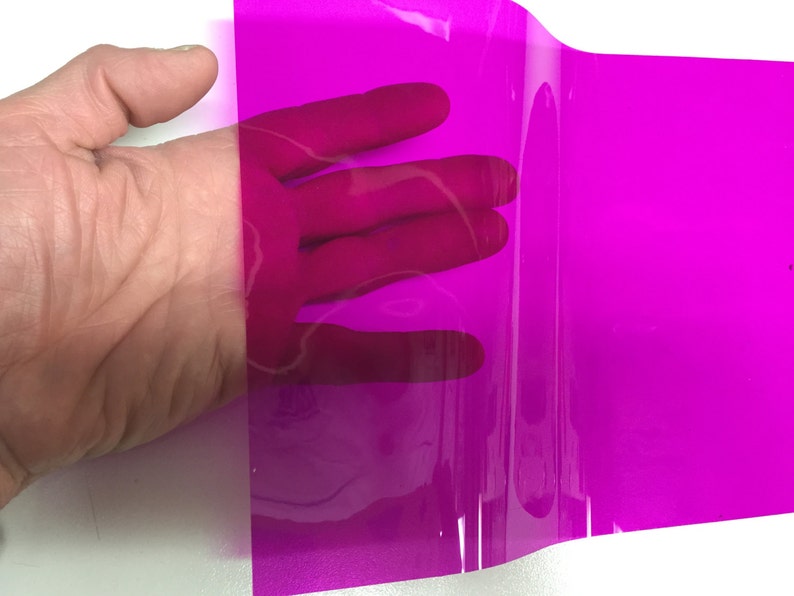 May include: A rectangular sheet of transparent magenta plastic film. The material is smooth, flexible, and semi-transparent, allowing light to pass through while casting a vibrant pink hue over the surface behind the sheet.