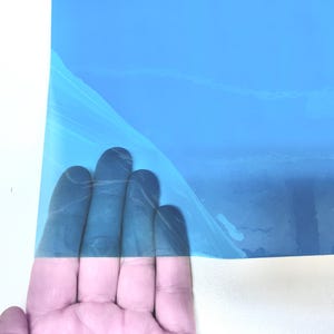 May include: A rectangular sheet of bright blue adhesive vinyl film with a protective backing layer partially peeled back to reveal the smooth, glossy surface of the material, showcasing its vibrant color and flexible, thin plastic texture.