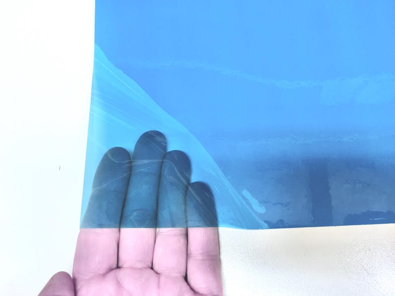 May include: A rectangular sheet of bright blue adhesive vinyl film with a protective backing layer partially peeled back to reveal the smooth, glossy surface of the material, showcasing its vibrant color and flexible, thin plastic texture.