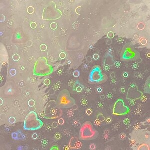 Puede incluir: Papel hologr&aacute;fico con un patr&oacute;n repetido de corazones, c&iacute;rculos y formas geom&eacute;tricas. Los corazones tienen un brillo de arco&iacute;ris, reflejando colores como verde, naranja y rosa. El fondo es de color claro y neutro.