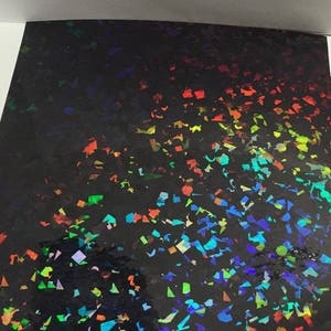 K&ouml;nnte beinhalten: Ein Bogen schwarzes holografisches Papier mit einem konfettiartigen Muster aus bunten, irisierenden Formen. Die Formen reflektieren einen Regenbogen aus Farben, darunter Rot, Orange, Gelb, Gr&uuml;n, Blau und Violett.