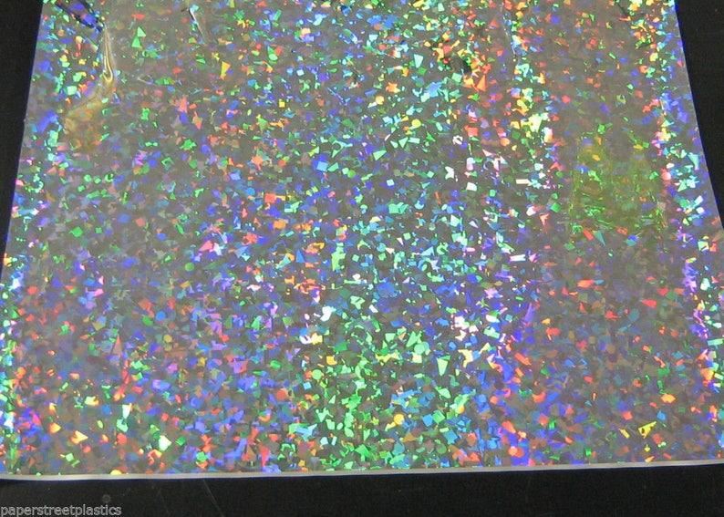 K&ouml;nnte beinhalten: Eine Folie aus schillerndem Kunststoff mit einem Streumuster aus bunten, unregelm&auml;&szlig;igen Formen. Die Farben umfassen Gr&uuml;n, Blau, Rot und Gelb und erzeugen einen schimmernden Effekt. Das Material ist reflektierend.