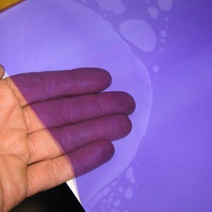 May include: A rectangular sheet of translucent purple film with a smooth texture. The material is semi-transparent, allowing the skin tone of the fingers placed underneath to be visible through the vibrant violet surface of the plastic sheet.
