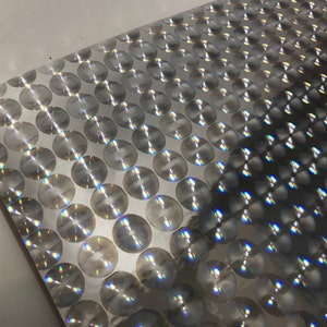 May include: A sheet of silver metallic material with a repeating pattern of circular, reflective discs. The discs have a textured, radial design, creating a shimmering effect. The material is slightly creased, showing its flexibility.