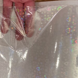 Puede incluir: L&aacute;mina de celof&aacute;n transparente e iridiscente con un patr&oacute;n repetido de corazones, c&iacute;rculos y copos de nieve. El dise&ntilde;o hologr&aacute;fico refleja un arco&iacute;ris de colores. El celof&aacute;n se sostiene, mostrando su flexibilidad y textura.