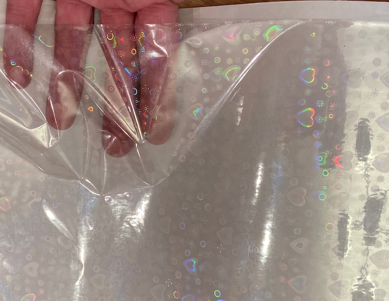 Puede incluir: L&aacute;mina de celof&aacute;n transparente e iridiscente con un patr&oacute;n repetido de corazones, c&iacute;rculos y copos de nieve. El dise&ntilde;o hologr&aacute;fico refleja un arco&iacute;ris de colores. El celof&aacute;n se sostiene, mostrando su flexibilidad y textura.