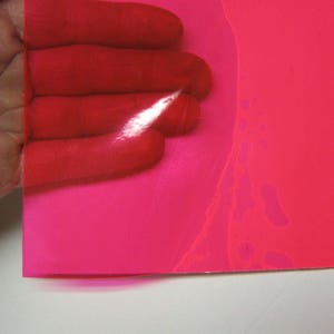 May include: A translucent, vibrant hot pink plastic sheet with a smooth, glossy finish. The material is flexible and semi-transparent, allowing light to pass through while casting a bright pink hue over the surface beneath it.