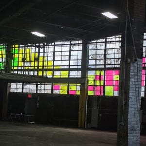 May include: A large industrial warehouse interior featuring expansive multi-pane windows. The glass panes are accented with vibrant, translucent squares in shades of neon pink, bright yellow, lime green, and orange, creating a colorful grid pattern.