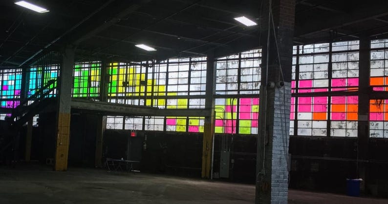 May include: A large industrial warehouse interior featuring expansive multi-pane windows. The glass panes are accented with vibrant, translucent squares in shades of neon pink, bright yellow, lime green, and orange, creating a colorful grid pattern.