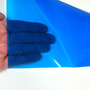 May include: A sheet of translucent, vibrant blue adhesive vinyl film. The material is smooth, glossy, and flexible, allowing light to pass through its surface. The blue film is partially peeled back from its white paper backing, revealing the adhesive side.