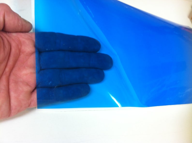 May include: A sheet of translucent, vibrant blue adhesive vinyl film. The material is smooth, glossy, and flexible, allowing light to pass through its surface. The blue film is partially peeled back from its white paper backing, revealing the adhesive side.