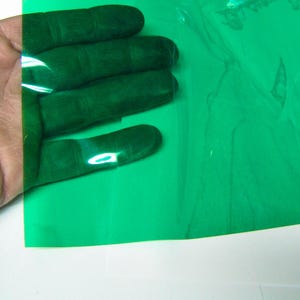May include: A flexible, translucent sheet of green plastic film. The material is smooth, glossy, and semi-transparent, allowing light to pass through while tinting the view with a vibrant green hue. The sheet is thin and pliable in texture.