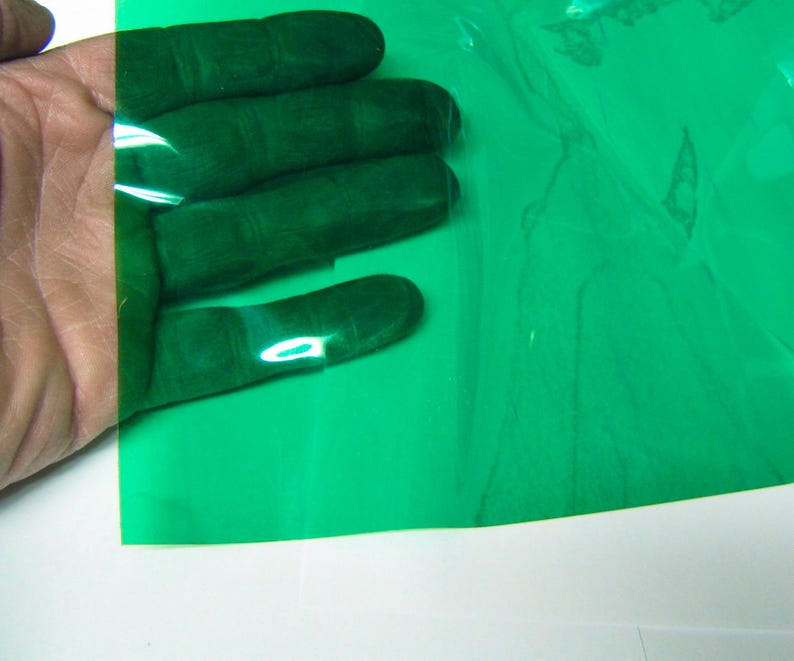 May include: A flexible, translucent sheet of green plastic film. The material is smooth, glossy, and semi-transparent, allowing light to pass through while tinting the view with a vibrant green hue. The sheet is thin and pliable in texture.