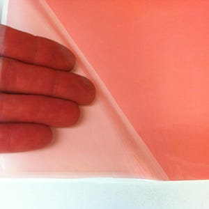 May include: A translucent, coral-colored adhesive film sheet with a partially peeled-back protective backing layer, revealing the smooth, semi-transparent surface of the material in a vibrant, solid orange-pink hue.