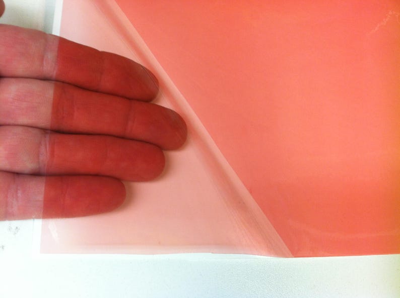 May include: A translucent, coral-colored adhesive film sheet with a partially peeled-back protective backing layer, revealing the smooth, semi-transparent surface of the material in a vibrant, solid orange-pink hue.