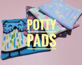Puede incluir: Una pila de almohadillas de entrenamiento coloridas con varios diseños, incluyendo una almohadilla azul con un diseño de gato y almohadillas con estampados florales y de conejos. El texto "POTTY PADS" está superpuesto en amarillo.