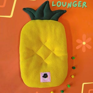 May include: A yellow pineapple-shaped lounger with a green leafy top. The lounger has a quilted pattern and a small pink label. The words "PINEAPPLE LOUNGER" are in the top right corner.