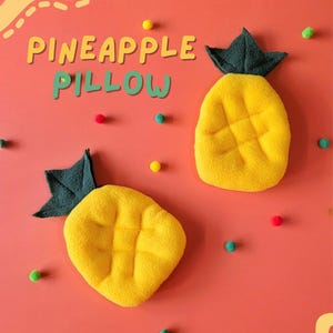 Peut inclure: Deux oreillers jaunes en forme d'ananas avec des dessus verts en forme de feuilles sur un fond corail. Les oreillers ont une surface texturée et sont entourés de pompons colorés. Les mots "PINEAPPLE PILLOW" sont écrits au-dessus des oreillers.
