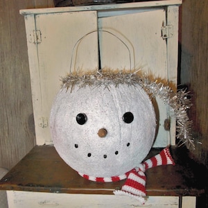 May include: A round white snowman bucket with a textured surface, featuring two black circular eyes, a small brown nose, and a dotted black smile. It has a silver wire handle, a silver tinsel trim, and a red and white striped knit scarf.