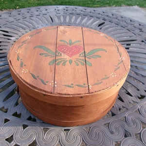 May include: A round, wooden box with a lid. The lid is decorated with a red heart and green leaves. The box is brown with a decorative floral design around the edge. The box is sitting on a metal table.
