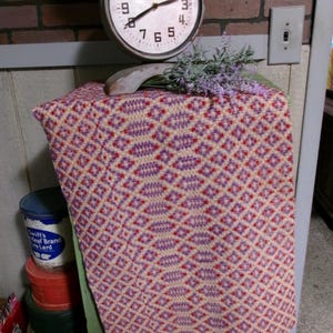 May include: A rectangular woven textile featuring a repeating geometric diamond pattern in shades of cream, purple, and red. The fabric is draped over a green wooden surface, positioned beneath a round analog clock with the text Antiques 1870.