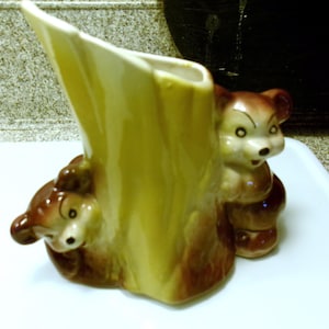 May include: A vintage ceramic vase featuring a tree trunk design with two brown bears peeking out. The vase has a light yellow interior and brown accents, creating a charming woodland theme. The bears have black eyes and a friendly expression.