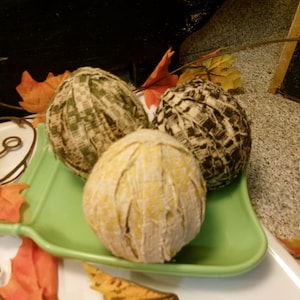 May include: Three spherical fabric-wrapped ornaments resting in a light green square dish. One sphere features a yellow and white pattern, another has a green and white checkered design, and the third displays a dark brown and white textured print.