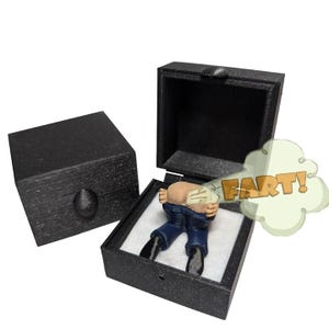 May include: A black square gift box containing a small figurine of a person bending over in blue jeans and black shoes. A cartoon-style graphic of a light green cloud with the orange text FART! extends from the figurine's rear.