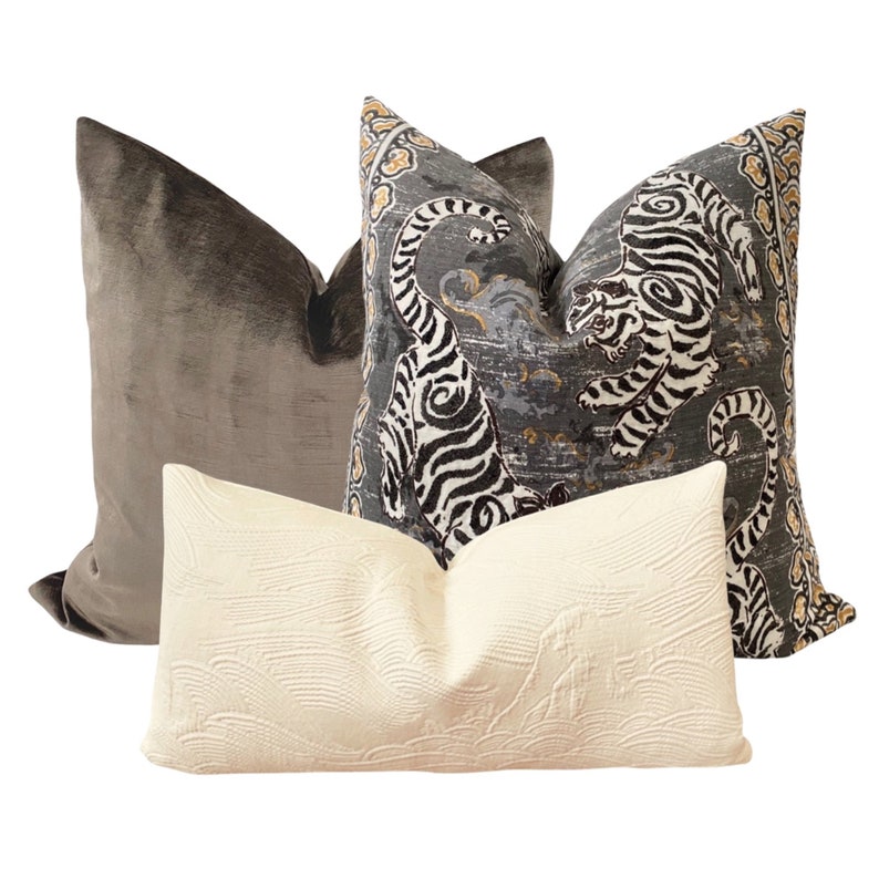 May include: Three decorative throw pillows. One pillow is a dark brown velvet, one is a cream colored pillow with a textured surface, and the third is a gray and black pillow with a tiger print.