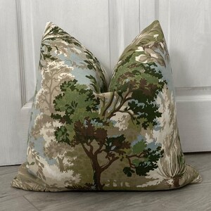 May include: A decorative throw pillow with a forest scene pattern. The pillow features a beige background with green, brown, and blue trees and foliage.