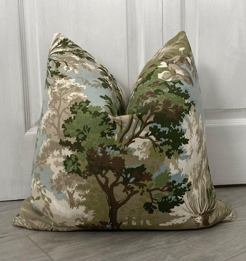 May include: A decorative throw pillow with a forest scene pattern. The pillow features a beige background with green, brown, and blue trees and foliage.