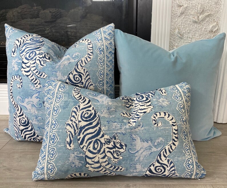 May include: Three decorative throw pillows with a blue and white tiger print. The pillows are arranged on a light wood floor. The pillow in the back is a solid light blue color.