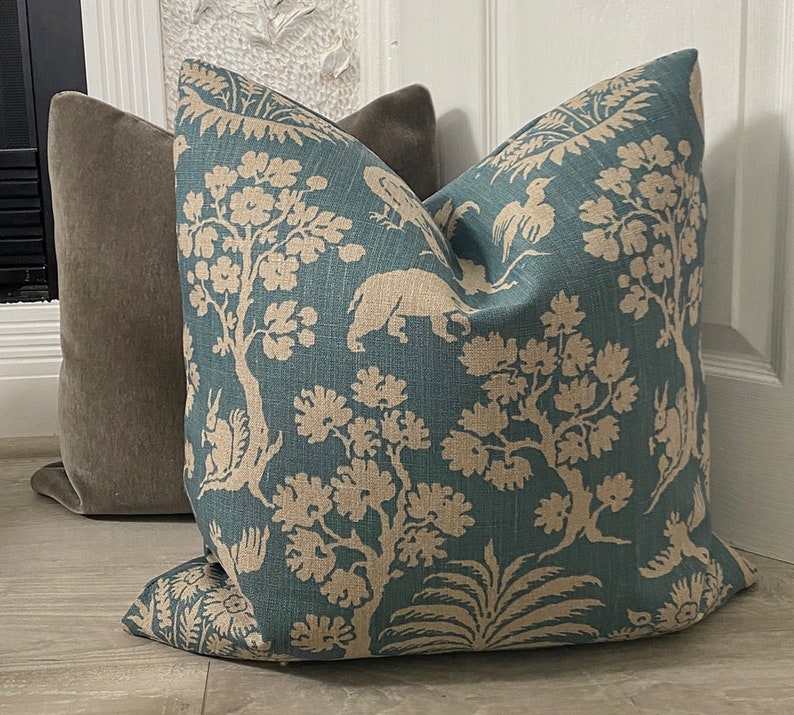 May include: A teal blue throw pillow with a beige floral and animal print. The pillow features a repeating pattern of trees, birds, and animals.