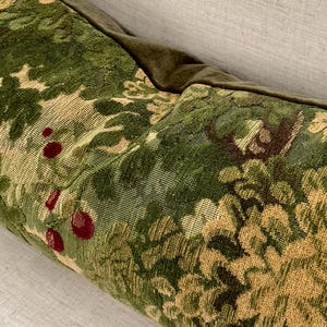 Puede incluir: Primer plano de un cojín decorativo con un estampado floral verde y beige. El cojín presenta un diseño de tapiz con acentos de bayas rojas. El borde superior es de terciopelo verde oscuro.