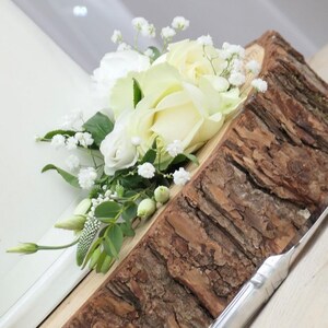 May include: White roses and baby's breath arranged on a rustic wooden slice. The flowers are arranged in a cluster and are partially visible. The wooden slice is a natural, rough-cut piece of wood with a bark edge.