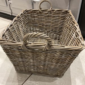 May include: A large, open-top wicker basket with two arched handles. The basket is woven in a light grey colour, with a rectangular shape and a slightly flared top. The basket is designed for storage and decorative purposes.