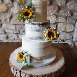 May include: A three-tier wedding cake with a rustic wood base. The cake is decorated with sunflowers and white flowers. The cake is a light colour with a subtle brown layer showing through.