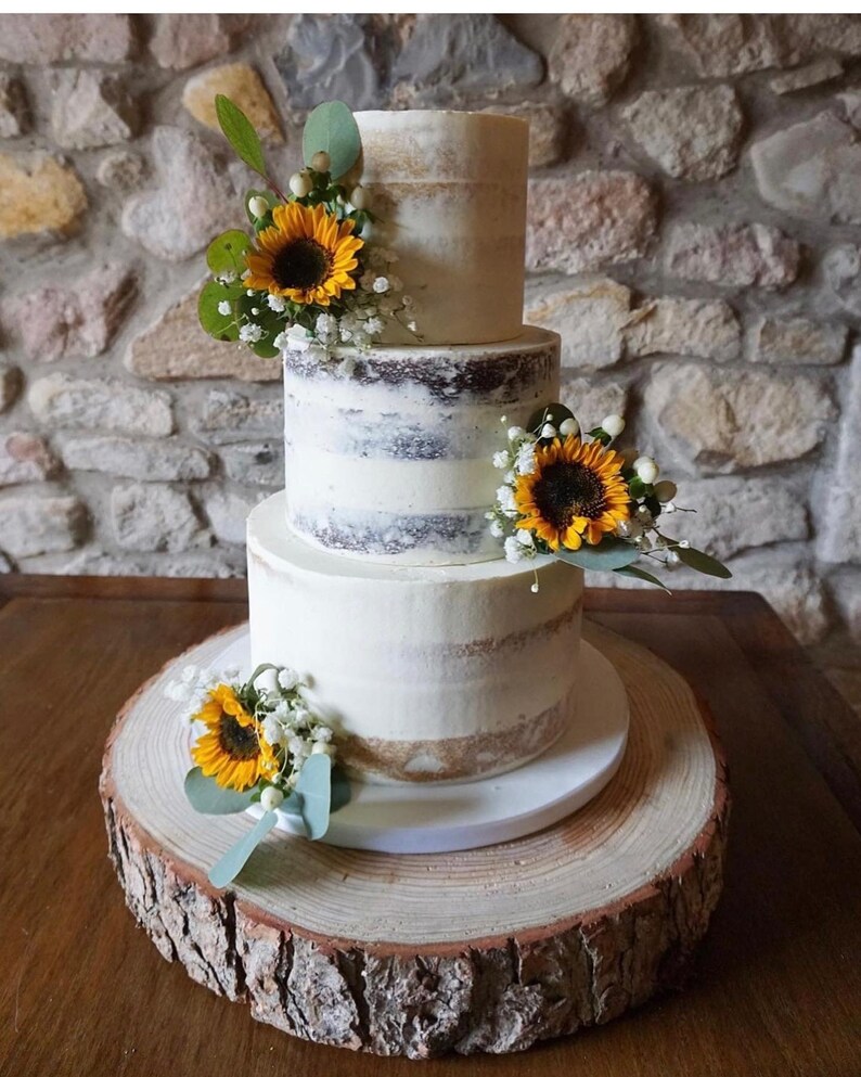 May include: A three-tier wedding cake with a rustic wood base. The cake is decorated with sunflowers and white flowers. The cake is a light colour with a subtle brown layer showing through.