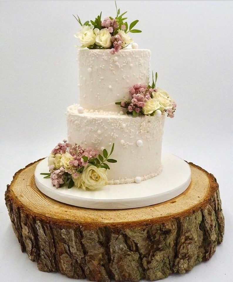 May include: Two-tiered white wedding cake decorated with white pearl sprinkles and fresh white and pink flowers on a wooden cake stand.