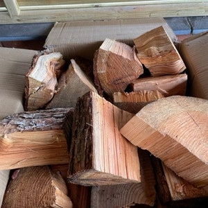 May include: A cardboard box filled with pieces of firewood. The wood is a light brown colour and has a rough texture. The box is sitting on a wooden surface.