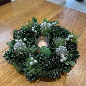 May include: A green pine cone wreath with silver berries and a small glass candle holder with a white candle in the centre.