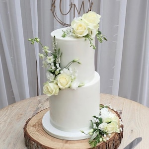 May include: A two-tiered white wedding cake decorated with white flowers and greenery. The cake is topped with a wooden cake topper with the initials "C & M" in a circle.