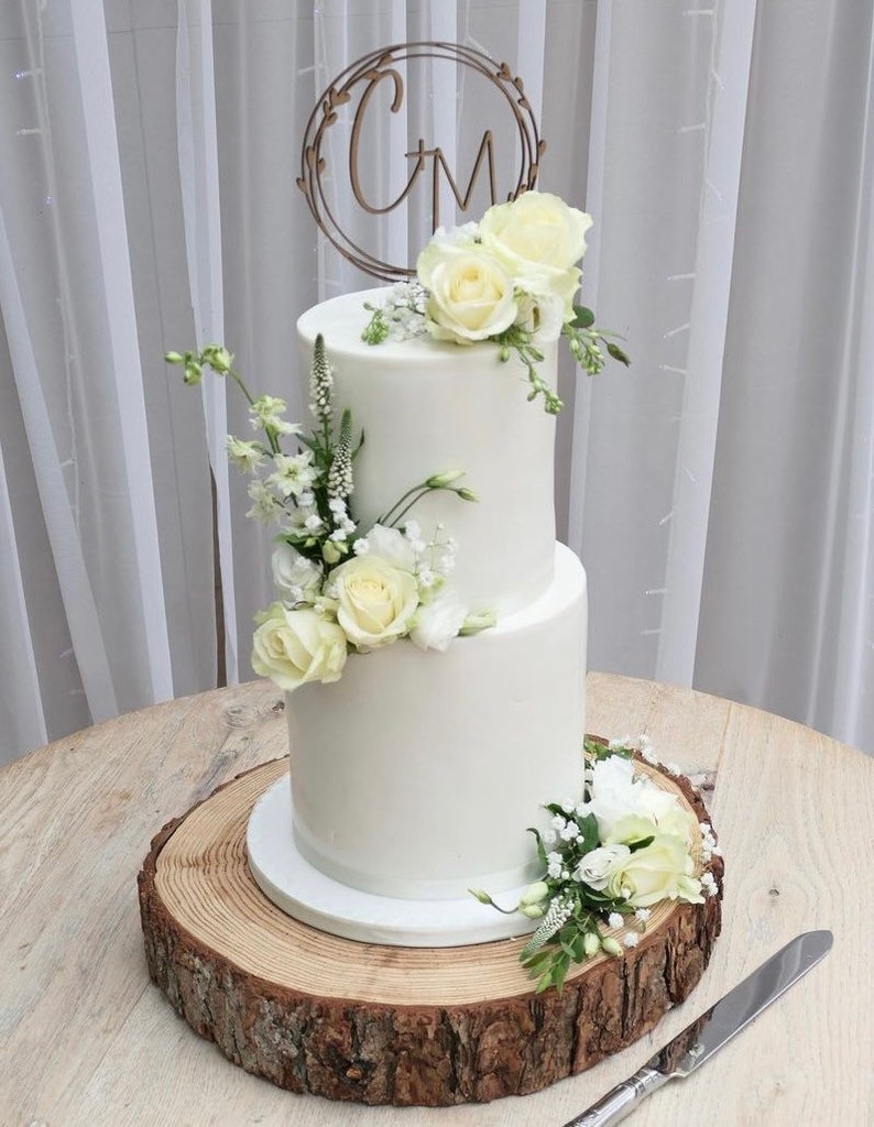 May include: A two-tiered white wedding cake decorated with white flowers and greenery. The cake is topped with a wooden cake topper with the initials "C & M" in a circle.