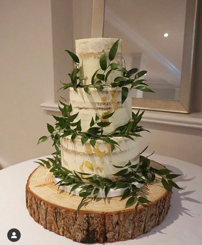 May include: A three-tier wedding cake with a rustic wooden base. The cake is decorated with greenery and gold leaf accents.