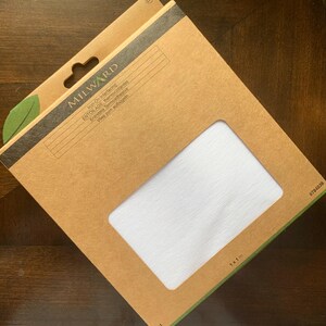 Pode incluir: Uma embalagem de entretela termocolante Milward. A embalagem de cartão castanho tem uma janela retangular que revela tecido branco. O texto na embalagem inclui "Iron-On Interfacing" e "1 x 1 m".