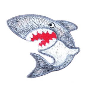 Può includere: Una toppa di squalo ricamata grigia con la bocca aperta rossa e un occhio bianco.
