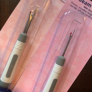 May include: Two seam rippers in clear plastic packaging. The rippers have a silver blade with a red safety ball and a white and grey handle. The packaging is pink with text that reads "Seam Ripper".