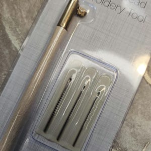 May include: A set of tambour bead embroidery tools, including a wooden handle with a gold metal hook and five needles in a clear plastic case. The packaging has the text "Tambour Bead Embroidery Tool" printed on it.