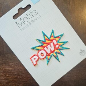 Può includere: Toppa da stirare o cucire con un motivo "POW!" giallo, rosso e blu. La toppa è su una carta bianca con il testo "Motifs Iron on & Sew on".