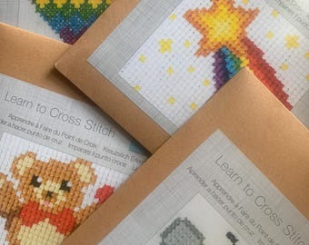 May include: Four Learn to Cross Stitch kits in brown paper envelopes. Each kit features a different cross-stitch pattern: a rainbow heart, a yellow star with a rainbow tail, a brown teddy bear, and a yellow and black bee on white fabric.